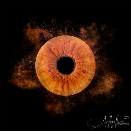 Un œil orange sur fond noir avec des teintes de poussière colorée autour.